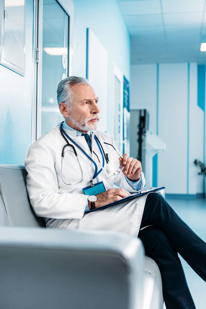 koncentruje się dojrzały mężczyzna lekarza ze schowka gospodarstwa, siedząc na kanapie w korytarzu szpitala  - Zdjęcie, obraz