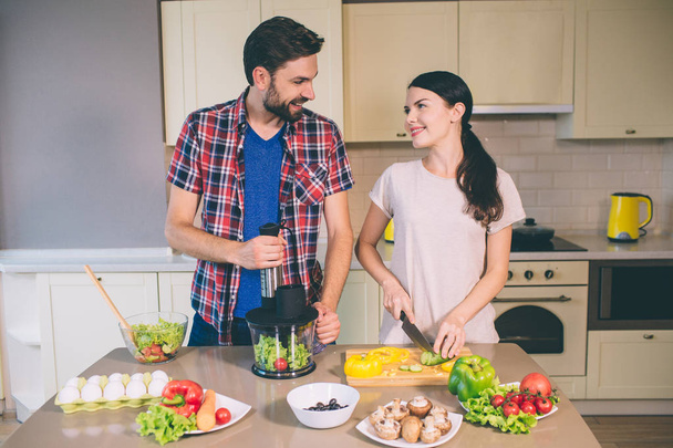 Delightful couple works together. Girl is cutting cucumber into pieces and looks at guy. He holds ingredients for salad in blender. Man looks at woman. They smile to each other. - Foto, Imagen