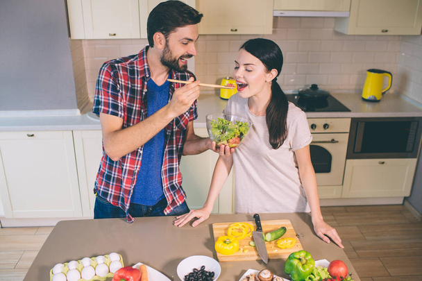 Cute picture of man standing at table and holding bowl with salad. He is stretching hand with wooden spoon with tomato on it. Girl opens her mouth. She wants to taste it. - Foto, Imagen
