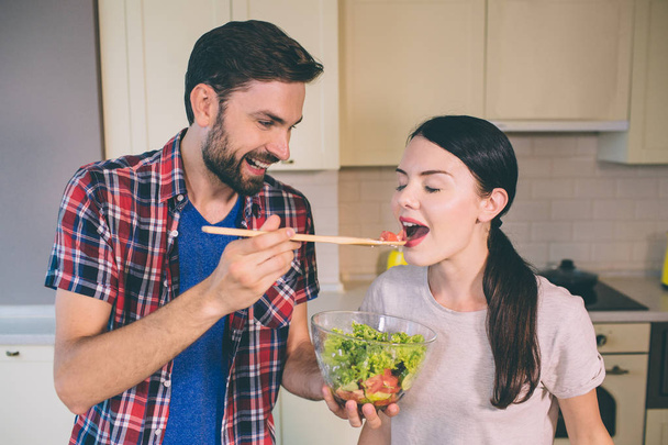 Horizontal portrait of couple standing at kitchen. Guy holds wooden spoon and feed girl. She keeps her mouth opened. They are tasing food they have cooked. - Photo, image