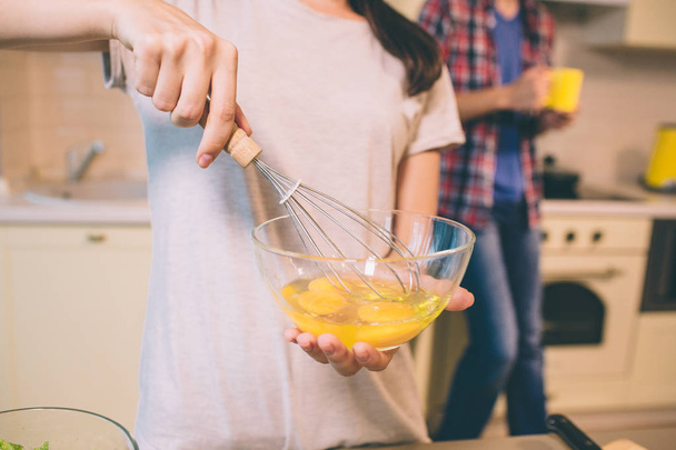Close up of girls hand blending eggs with special equipment in glass bowl. Guy stands behind her and holds yellow cup. - Photo, image