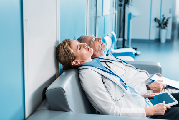 クリップボードと病院の廊下のソファーに空白の画面を持つデジタル タブレット寝て疲れている男性と女性医師の側面図  - 写真・画像