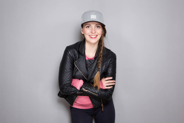 Portrait of cool young woman with leather jacket and hat standing by gray wall - Photo, Image