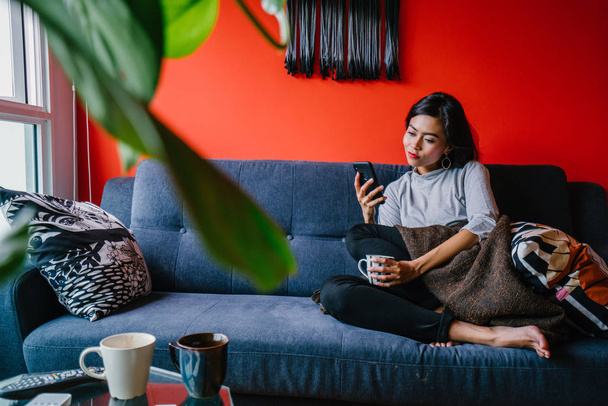 Portrait of a Singaporean Malay woman sitting on a comfortable blue couch with phone in a trendy living room during the daytime. She is relaxing and chilling out on her own on a weekend.  - 写真・画像