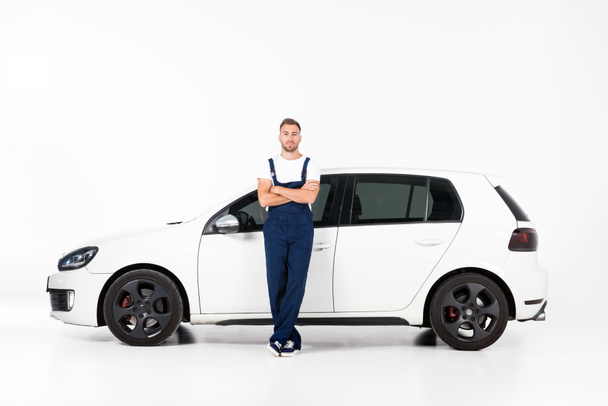 handsome auto mechanic leaning on car with crossed arms and looking at camera on white - Foto, afbeelding