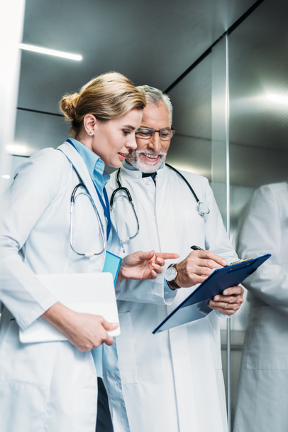 Улыбающийся взрослый врач-мужчина показывает планшет коллеге-женщине с цифровым планшетом в больничном лифте
  - Фото, изображение