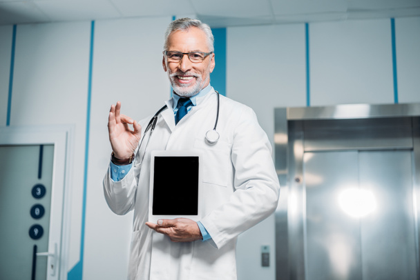 smiling mature male doctor with stethoscope over neck doing ok gesture and showing digital tablet with blank screen in hospital  - Photo, Image