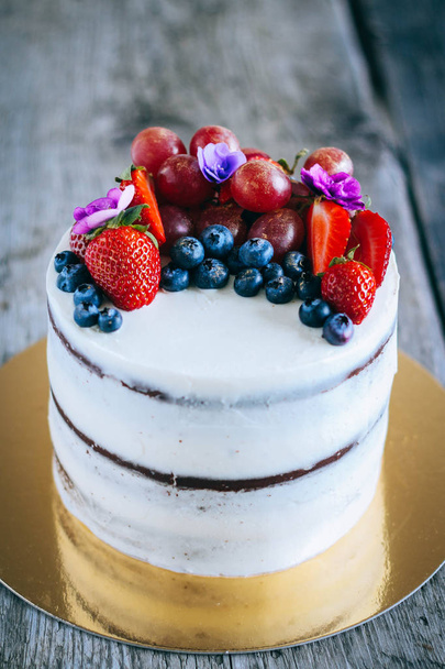 Chocolate cake with creamy cream decorated with strawberries, blueberries, grapes and violets on a wooden background - Photo, Image