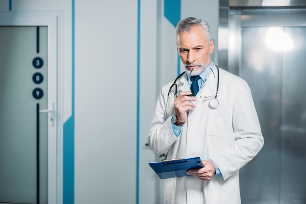pensive mature male doctor holding eyeglasses and clipboard in hospital  - Photo, Image