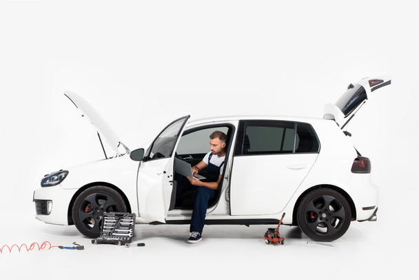handsome auto mechanic sitting in broken car and using laptop on white - Foto, Bild