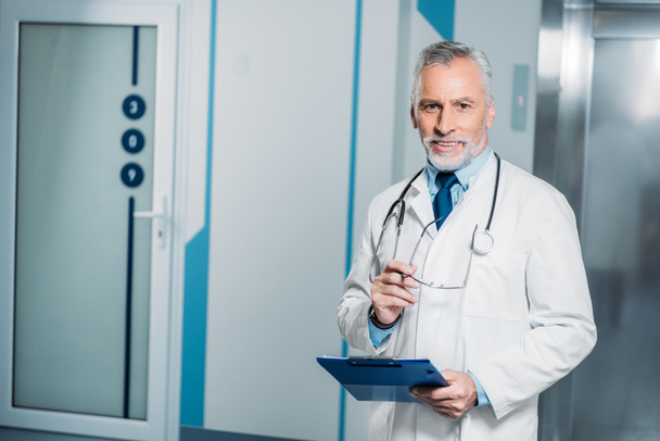 uśmiechający się dojrzały mężczyzna lekarza z stetoskop nad szyją trzymając okulary ze schowka i patrząc na kamery w szpitalu  - Zdjęcie, obraz