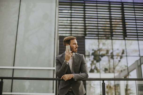 junger erfolgreicher Geschäftsmann trägt grauen Anzug und hält sein Smartphone in der Nähe moderner Büros oder Wolkenkratzer - Foto, Bild