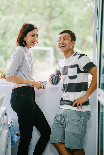 Young Malay Asian couple enjoying a hot drink and talking to one another fondly. They are sharing a tender moment together during the day by the window balcony. - Foto, imagen