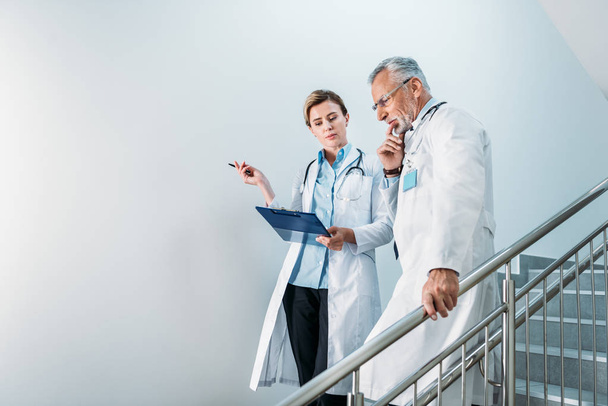 adulte femme médecin montrant presse-papiers à réfléchi collègue masculin sur escalier à l'hôpital
  - Photo, image