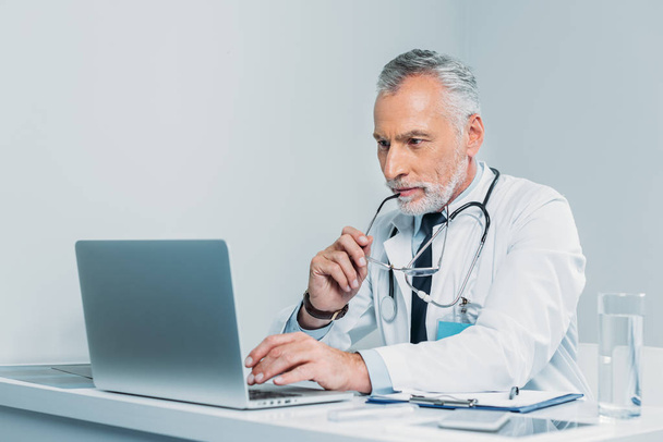 middle aged male doctor holding eyeglasses and using laptop at table in office  - Φωτογραφία, εικόνα