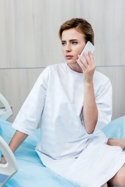 sad female patient looking away and talking on smartphone in hospital room - Photo, Image