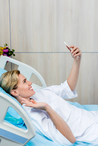 笑顔で手を振っていて、病室でスマート フォンでビデオ通話のメスの患者  - 写真・画像