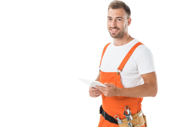 portrait of smiling handsome auto mechanic in orange uniform holding tablet isolated on white - Foto, Imagem