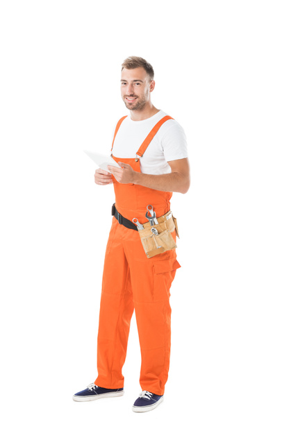 smiling handsome auto mechanic in orange uniform holding tablet isolated on white - Valokuva, kuva