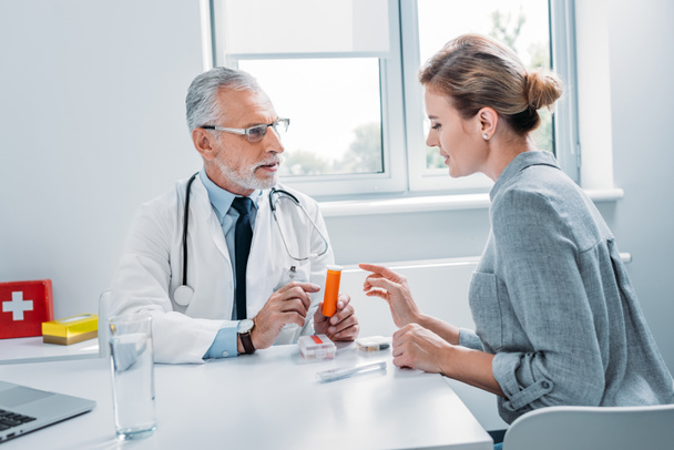 médico masculino de mediana edad señalando píldoras a paciente femenino en la mesa en el consultorio
  - Foto, Imagen