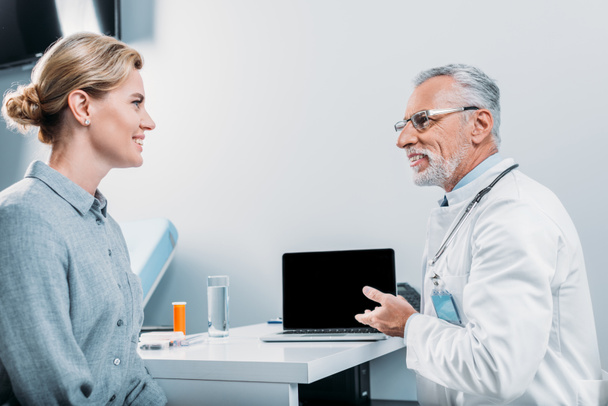 doctor masculino maduro feliz que señala el ordenador portátil con la pantalla en blanco al paciente femenino que se sienta cerca en la oficina
  - Foto, Imagen