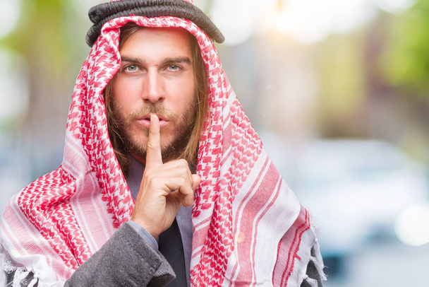 Genç yakışıklı Arap adam uzun saçlı dudaklarında parmak ile sessiz olmanı izole arka plan üzerinde keffiyeh giyiyor. Sessizlik ve gizli kavramı. - Fotoğraf, Görsel