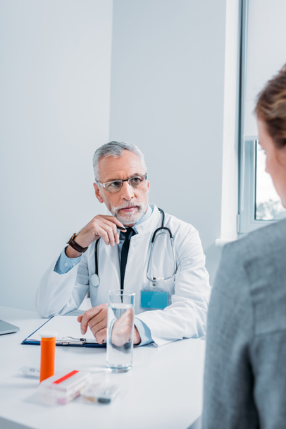 serious mature male doctor talking to female patient at table with pills and clipboard in office  - Foto, Imagen