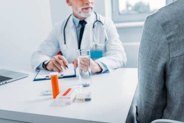 錠剤や office のクリップボードとのテーブルで女性患者に話している男性医師の部分的なビュー  - 写真・画像