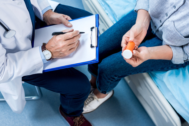 imagen recortada de médico varón maduro escribiendo en portapapeles cerca de paciente femenino con pastillas en la habitación del hospital
 - Foto, Imagen