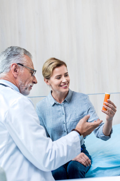 glimlachend volwassen mannelijke dokter geven pillen aan vrouwelijke patiënt in ziekenhuis kamer - Foto, afbeelding