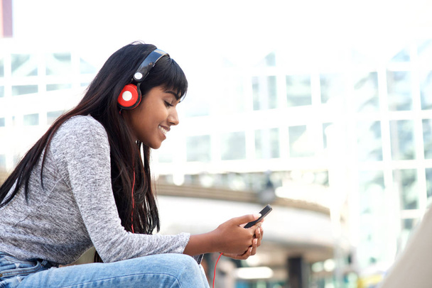 Portret stronie uśmiechający się młodej kobiety Indian siedział zewnątrz w mieście słuchanie muzyki z telefonu komórkowego i słuchawki - Zdjęcie, obraz