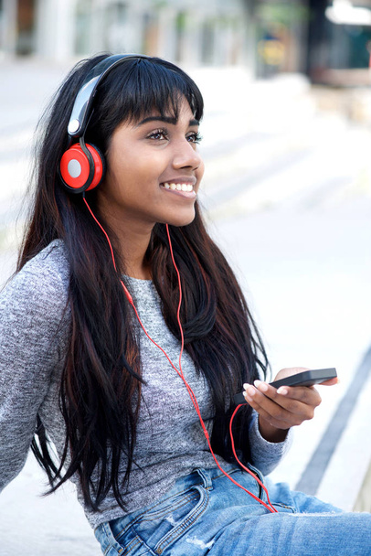 Портрет улыбающейся молодой индийской женщины, сидящей снаружи с мобильным телефоном и наушниками
 - Фото, изображение