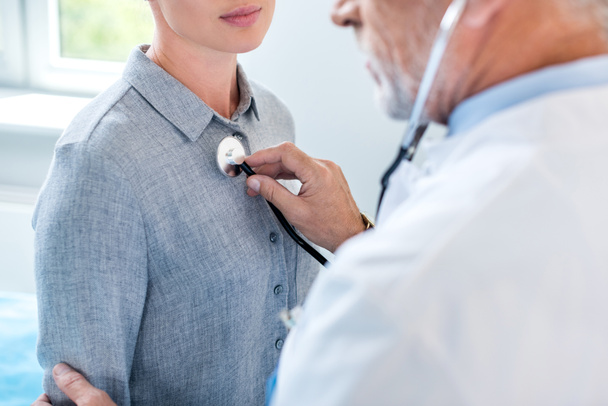 Teilansicht eines reifen männlichen Arztes, der Patientin im Krankenhauszimmer mit dem Stethoskop untersucht - Foto, Bild
