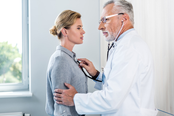 boční pohled na zralý muž Doktor vyšetřují pacientky o stetoskop v nemocničním pokoji - Fotografie, Obrázek