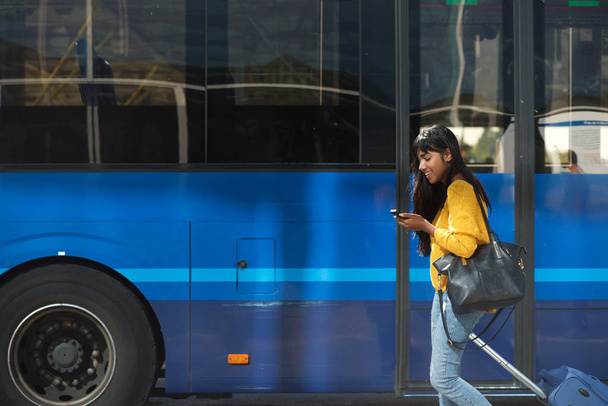 Side portrait of smiling young Indian woman walking with suitcase and cellphone by bus station - Photo, image