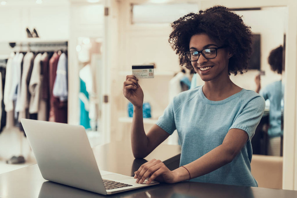 Young Black Woman holding Credit Card in Store. Portrait of Happy Beautiful African Girl holding Credit Card while using Laptop in Boutique inside Mall. Female Employee takes Payment from Customer - Valokuva, kuva