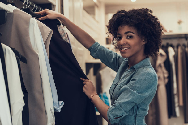 Smiling Young Woman Shopping in Clothing Store. Happy Beautiful Black Girl choosing Clothes to Buy in Boutique inside Mall. Attractive african Woman with Afro hairstyle in Clothing Store - Foto, Bild