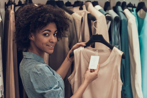 Smiling Young Woman Shopping in Clothing Store. Happy Beautiful Black Girl choosing Clothes to Buy in Boutique inside Mall. Attractive african Woman with Afro hairstyle in Clothing Store - Zdjęcie, obraz