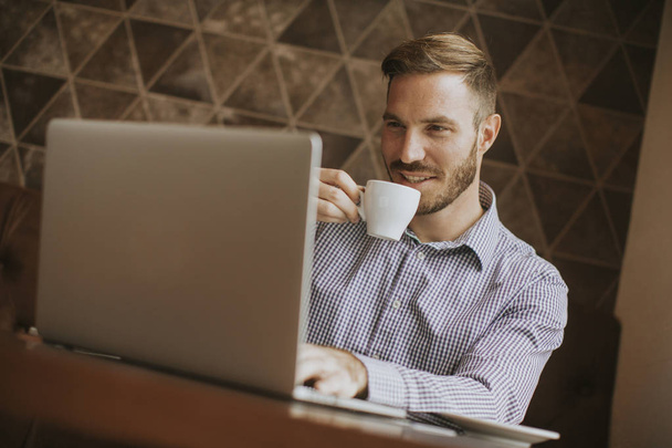 Вид на молодого человека, сидящего в кафе, пьющего кофе и с улыбкой смотрящего на ноутбук
 - Фото, изображение