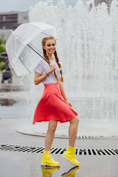 Young pretty girl with two braids in yellow boots and with transparent umbrella stands near fountain. Rainy day in city. - Фото, изображение
