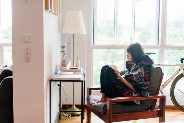 A young Malay Asian woman sitting  and working  on her computer at home in a corner in her study or living room during the day. She is comfortably on a chair with her legs propped up. - Foto, imagen