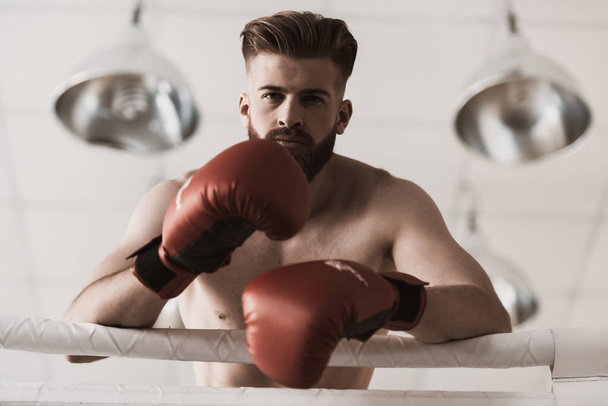 Portret van een mannelijke bokser in handschoenen op de boksring. Portret van een man met een baard die rode bokshandschoenen draagt... staand op een ring leunend op een touw voor het gevecht. Sterkte en motivatie Concept - Foto, afbeelding