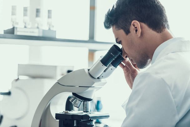 Young Scientist using Microscope in Laboratory. Male Researcher wearing white Coat sitting at Desk and looking at Samples by using Microscope in Lab. Scientist at Work in Laboratory - 写真・画像