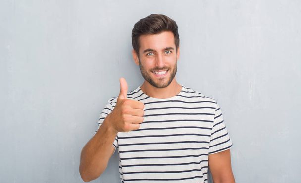 Handsome young man over grey grunge wall wearing navy t-shirt doing happy thumbs up gesture with hand. Approving expression looking at the camera with showing success. - Zdjęcie, obraz