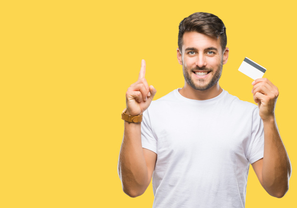 Young handsome man holding credit card over isolated background surprised with an idea or question pointing finger with happy face, number one - Foto, Bild