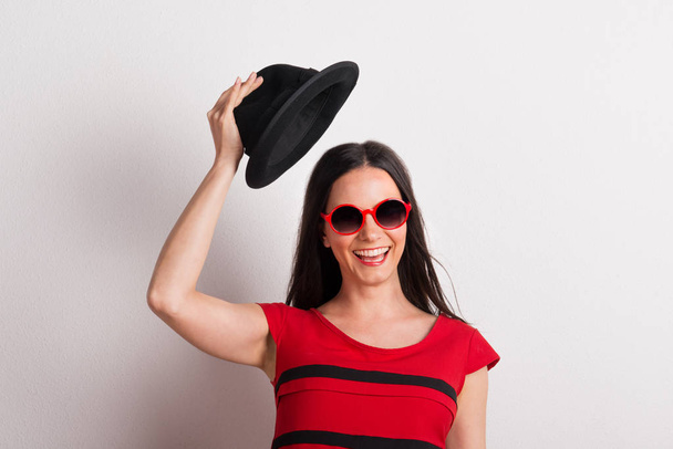 A young beautiful woman with red sunglasses lifting black hat in a studio. - Fotoğraf, Görsel