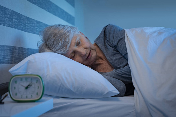 Old woman in grey hair sleeping peacefully at night time in bed. Senior woman lying on side and sleeping at home. Mature woman feeling relaxed at home while sleeping at night. - Foto, imagen