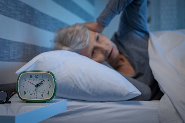 Old woman suffering from migraine unable to sleep during the late night. Elderly woman in bed ill and suffering from sleeplessness and insomnia. Closeup of alarm clock showing time with senior woman unable to sleep in background.  - Foto, immagini