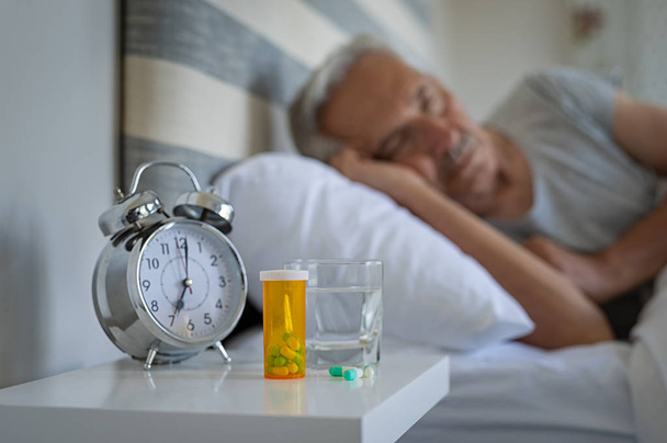 Medical pills and alarm clock with senior man sleeping in background. Silver alarm clock and medicines on table while man resting on bed. Bottle of capsules with glass of water kept on night table. - Photo, image