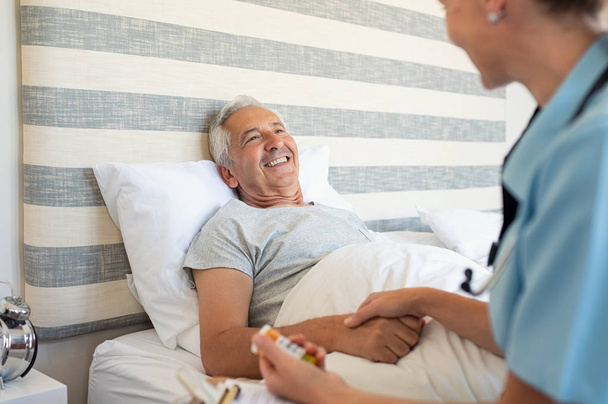Cheerful senior man lying on bed receiving health care at home. Nurse advising old man on medication at home. Caregiver nurse helping elderly man taking medicine on the bed - Foto, Imagen